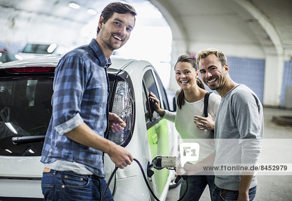 Porträt von glücklichen Freunden mit einem Mann  der an der Tankstelle ein Elektroauto auflädt.