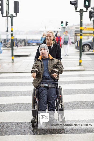 Hausmeister schiebt behinderten Mann im Rollstuhl  während er die Straße überquert.
