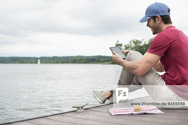 A man seated on a jetty by a lake  using a digital tablet.