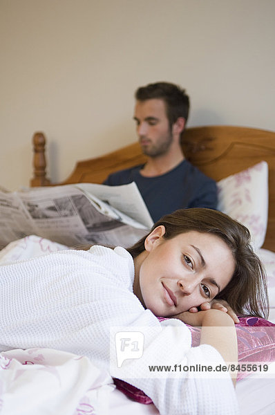 A man reading the paper in bed  and a woman relaxing smiling.