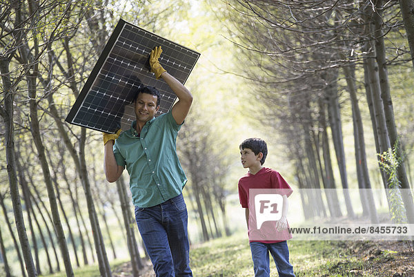 Ein Mann  der einen Sonnenkollektor eine Baumallee hinunterträgt  begleitet von einem Kind.