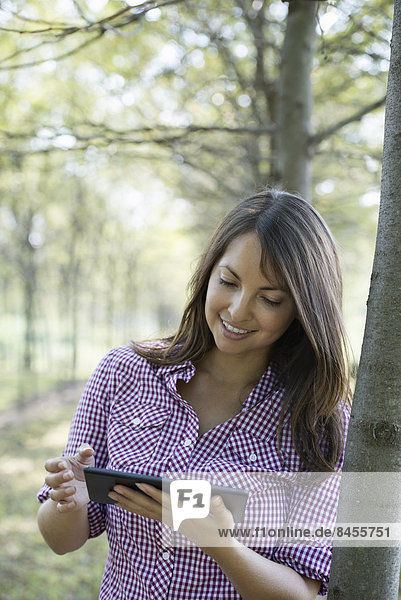 Eine Frau steht in einer Baumallee und hält ein digitales Tablett in der Hand.