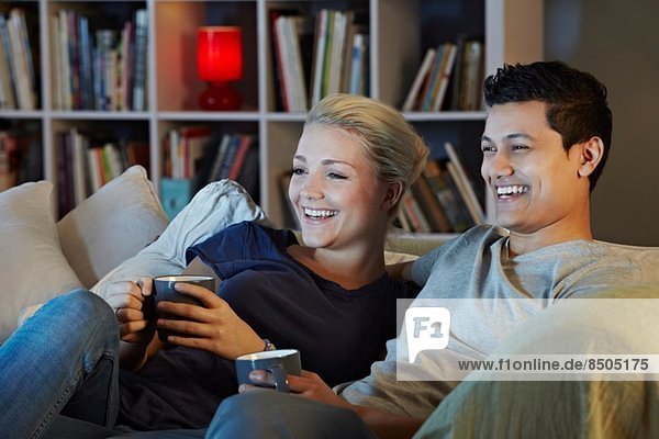 Young couple sitting on sofa with hot drinks  watching tv