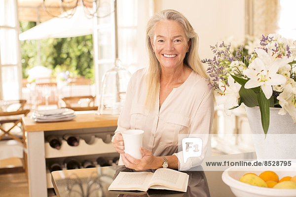 Senior woman drinking coffee and reading book in kitchen