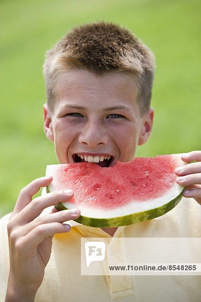 A teenage boy taking a large bite out of a watermelon.