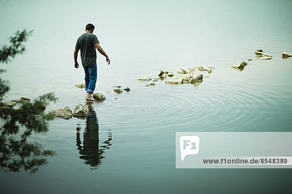 Ein Mann  der barfuß über Trittsteine vom Ufer eines Sees weggeht.