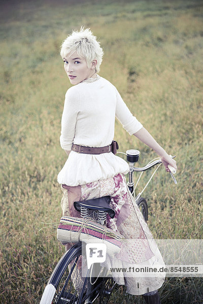 Ein Mädchen in Vintage-Kleidung Baumwollrock mit Paisleymuster und ein Fahrrad im langen Gras.