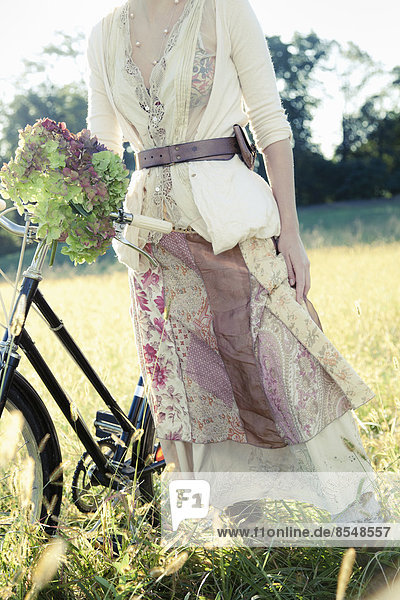 Ein Mädchen in Vintage-Kleidung Baumwollrock mit Paisleymuster und ein Fahrrad im langen Gras.