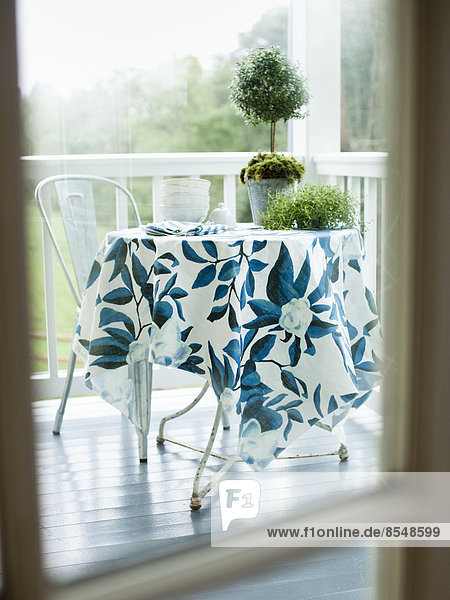 Ein runder Tisch und Stuhl aus Schmiedeeisen auf einer weiß gestrichenen Hausveranda  mit einer blau gemusterten Tischdecke.