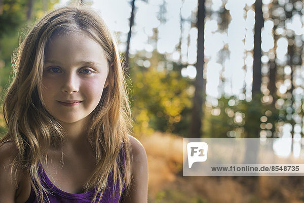 A young girl with long blonde hair in woodland in the fresh air  looking at the camera.