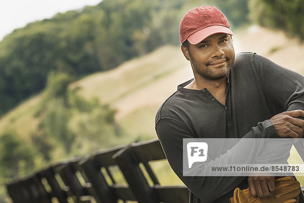 Ein junger Mann mit einer Baseballmütze  der auf einem Gehweg mit ländlichem Hintergrund steht.