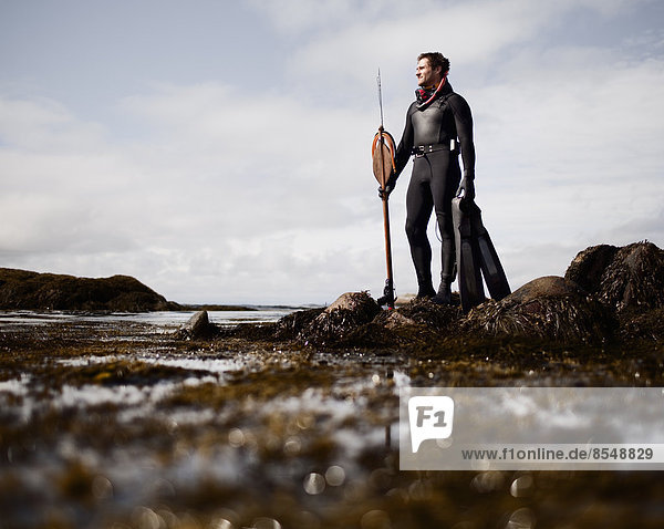 A man in a wetsuit  standing on the shore with a large spear fishing harpoon.