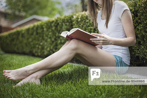 Ein Mädchen sitzt auf dem Rasen in einem Garten und liest ein Buch.