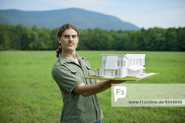 Eine ländliche Szene und eine Bergkette sowie eine Person  die ein maßstabsgetreues Modell eines neuen Gebäudes hält.