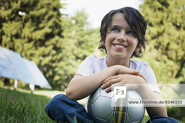 Ein Junge  der einen Fußball hält und auf dem Rasen sitzt. Sonnenkollektoren.