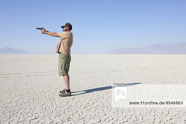 A man aiming a hand gun  holding it with his arm outstretched  standing in a vast  barren desert.