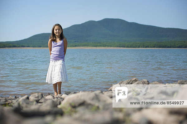 A young girl in a white skirt and purple top. Standing on the shore of a lake.