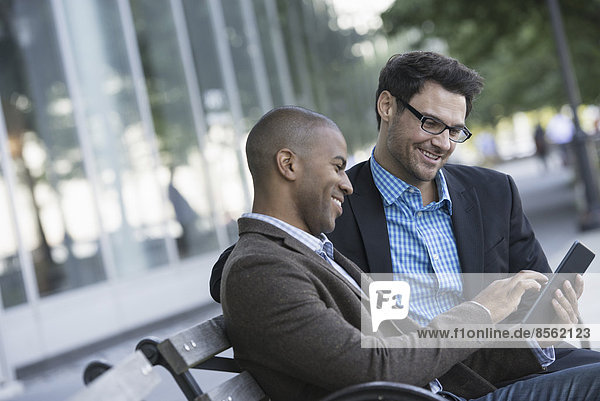 Business people in the city. Keeping in touch on the move. Two men seated on a park bench outdoors  looking at a digital tablet.
