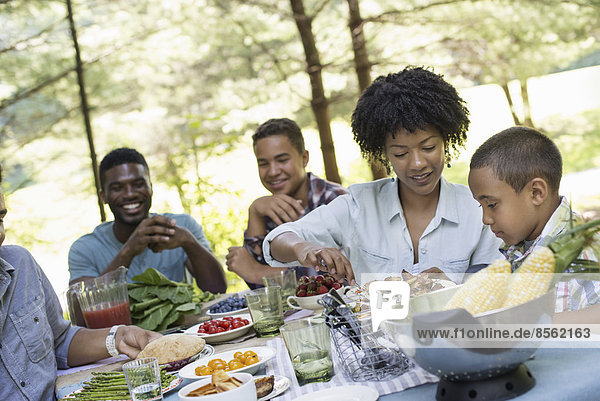 A family picnic in a shady woodland. Adults and children around a table handing around plates and food.