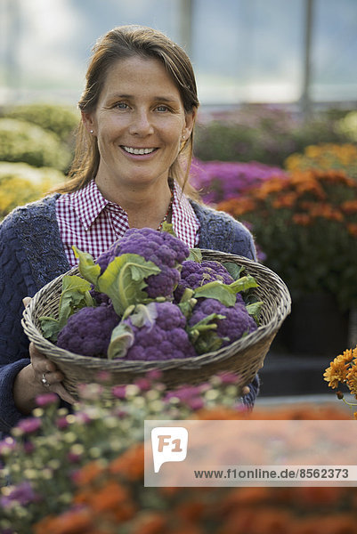 Eine Frau hält eine Schüssel mit frischem Obst und Gemüse in der Hand  aus dem violetter Brokkoli sprießt. Blühende Pflanzen. Krysanthemen.
