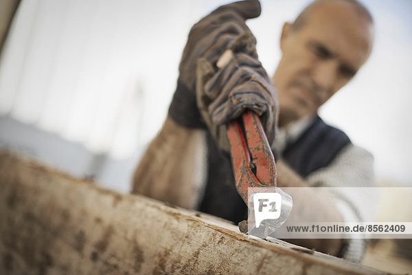 Ein Mann arbeitet auf einem ehemaligen Holzlager. Er benutzt ein Werkzeug  um Metalle aus einem Stück Altholz zu entfernen.