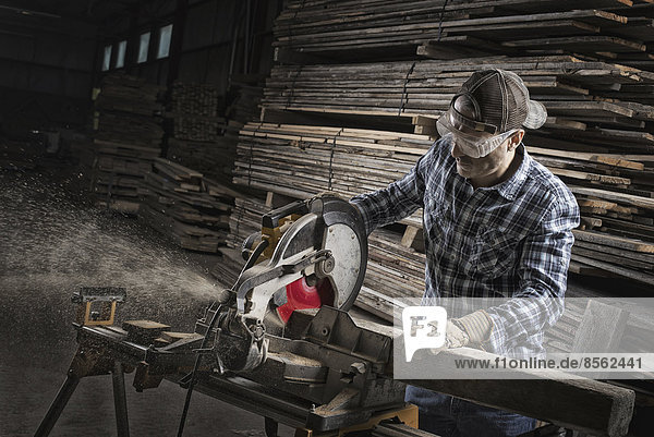 Eine zurückgewonnene Holzwerkstatt. Ein Mann mit Schutzbrille  der mit einer Kreissäge Holz sägt.