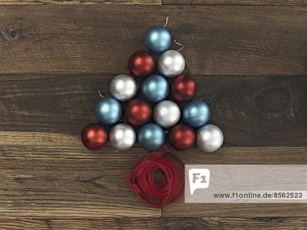 Eine Sammlung blauer,  roter und silberner Ornamente,  die in Dreiecksform auf einem Holzbrett angeordnet sind. Eine Weihnachtsbaumform mit einem roten Band,  das an der Basis aufgewickelt ist.