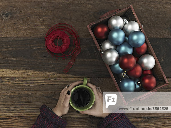 Eine Sammlung von blauen  roten und silbernen Ornamenten und rotem Band in einer Kiste auf einem Holzbrett. Eine um eine Tasse Kaffee geschlungene Hand. Eine Pause machen.