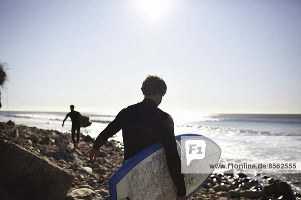 Zwei Personen in Neoprenanzügen an einem Strand  die ihre Surfbretter tragen.