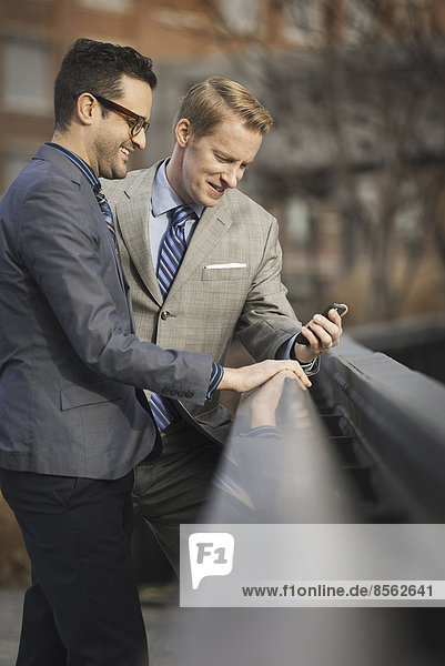 Zwei Männer in formeller Geschäftskleidung  die Seite an Seite stehen und auf einen Handybildschirm oder ein Mobiltelefon schauen.