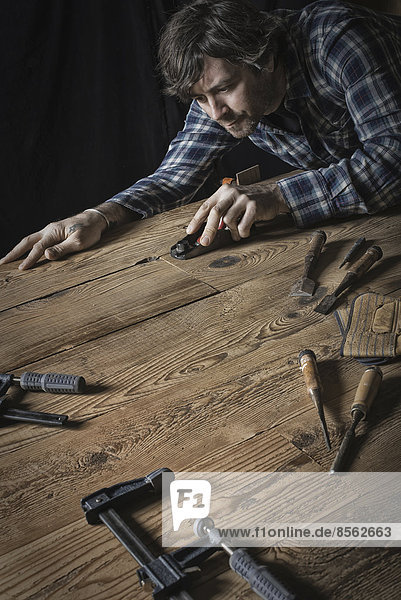 A man working in a reclaimed lumber yard workshop. Holding tools and sanding knotted and uneven piece of wood.