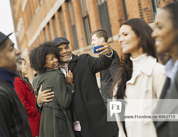 Stadtleben. Eine Gruppe von Menschen unterwegs. Ein Mann hält ein Fotohandy in die Hand und macht Fotos von der Gruppe. Ein Mann küsst eine junge Frau. Männer und Frauen.