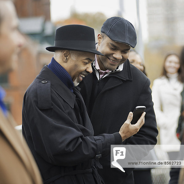 Stadtleben. Zwei Männer stehen Seite an Seite  halten Kontakt  benutzen Mobiltelefone und schauen lachend auf den Bildschirm.