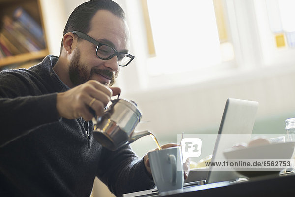 A bearded man having a drink of coffee. An open laptop computer on the counter.
