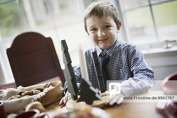 Kinder in einem Familienhaus. Ein kleiner Junge an einem Tisch  neben einem großen Tablett mit Keksen. Zwei hölzerne Christbaumschmuckstücke.