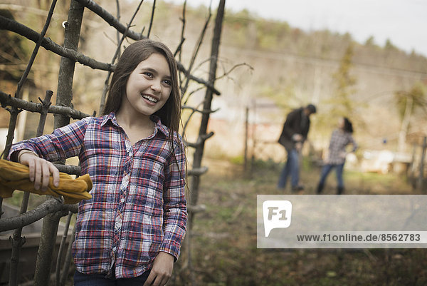 Ein junges Mädchen  das sich an einen Zaun lehnt  und zwei Personen im Hintergrund. Ein Bio-Bauernhof.