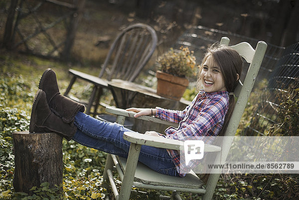A young girl sitting in a wooden chair in a garden  with her feet up on a log.