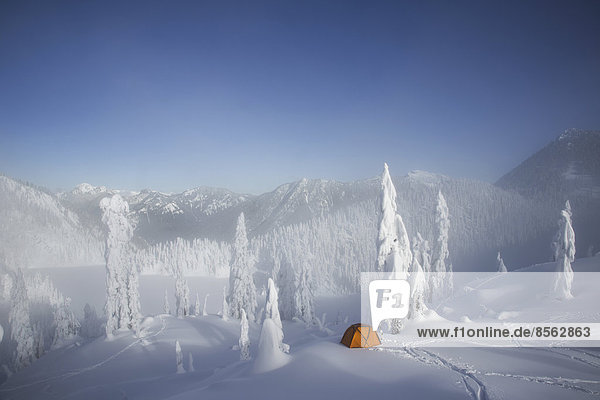 Ein leuchtend orangefarbenes Zelt zwischen schneebedeckten Bäumen,  auf einem verschneiten Bergrücken mit Blick auf einen Berg in der Ferne.