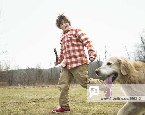 A young boy outdoors on a winter day  holding a stick and running with a golden retriever dog.