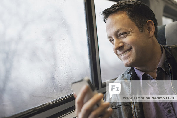 Ein reifer Mann  der an einem Fensterplatz in einem Zug sitzt und sein Mobiltelefon in der Hand hält. Er lächelt und schaut in die Ferne.