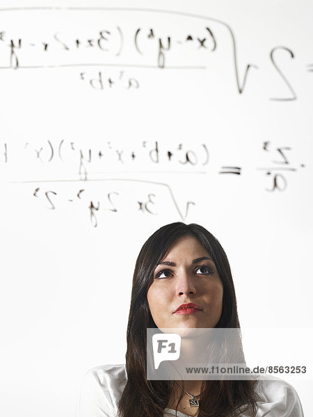 Eine junge Frau  die eine mathematische Gleichung mit schwarzem Marker auf eine durchsichtige  durchsichtige Fläche schreibt und sich zurücklehnt  um sie zu betrachten.