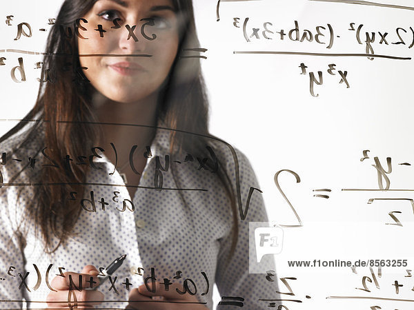 Eine junge Frau betrachtet eine mathematische Gleichung  die mit einem schwarzen Marker auf eine durchsichtige  durchsichtige Wand geschrieben wurde.