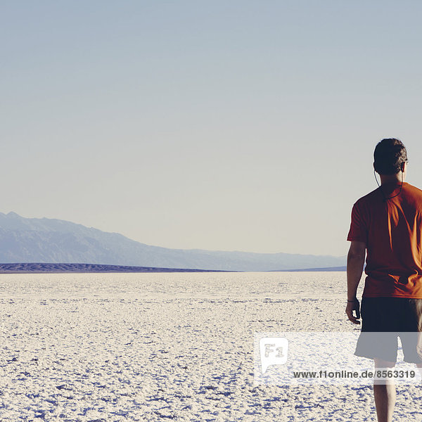 A man in a red shirt and shorts  standing on the salt flats overlooking the desert at Badwater  Death Valley national park.