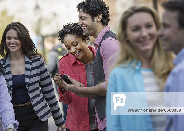 People outdoors in the city in spring time. A group of men and women  two looking at a cell phone screen and laughing.