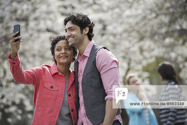 Menschen im Frühling in der Stadt im Freien. Weiße Blüte an den Bäumen. Eine junge Frau macht ein Foto von sich und einem jungen Mann.