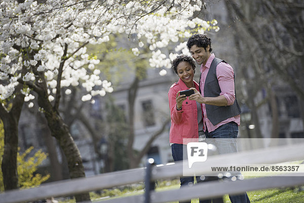 People outdoors in the city in spring time. New York City park. A man and woman side by side. Taking a photograph with a hand held cell phone.