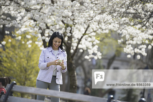 Draußen in der Stadt im Frühling. Park von New York City. Weiße Blüte an den Bäumen. Eine Frau hält ihr Handy in der Hand und lächelt.