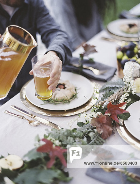 Eine Gruppe von Menschen um einen Tisch in einem Garten. Ein Festmahl mit gedecktem Tisch und Blattdekoration. Eine Person  die Getränke in Gläser gießt.