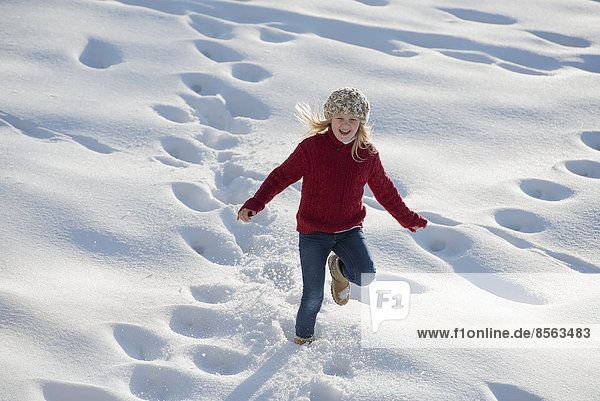 Winterlandschaft mit Schnee auf dem Boden. Ein junges Mädchen rennt durch Tiefschnee und hinterlässt Fußspuren.