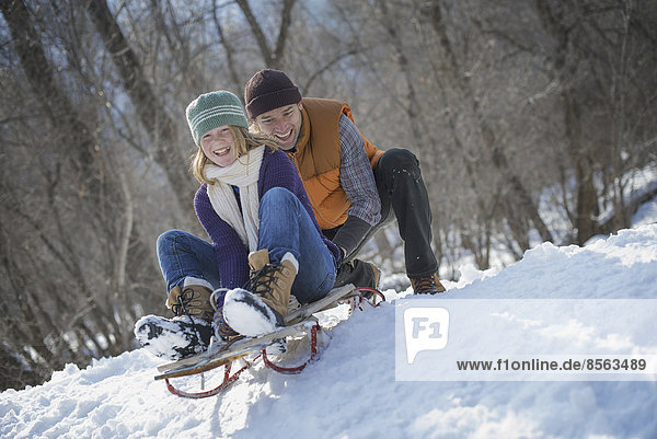 Winter scenery with snow on the ground. A man pushing a young woman from the top of a slope on a toboggan.